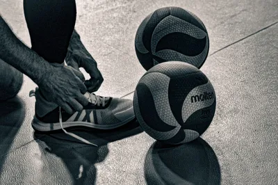 Photo noir et blanc en plongée : un joueur assis noue ses chaussures, deux ballons posés au sol à côté de lui.