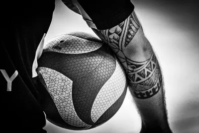 Photo noir et blanc en gros plan : un ballon calé contre le bras tatoué d'un joueur, motifs polynésiens visibles.